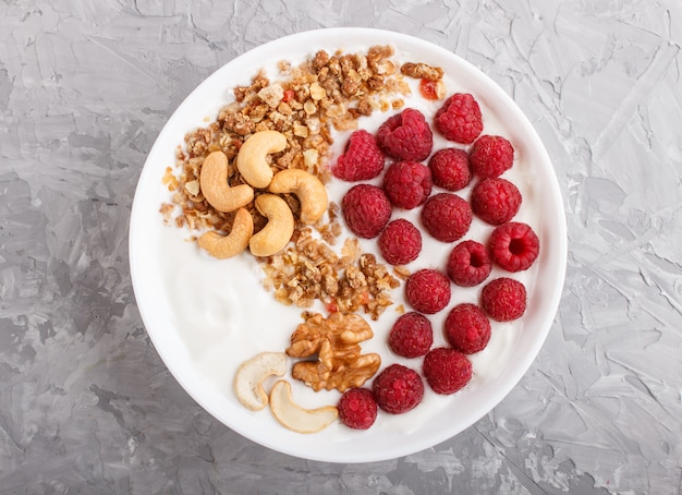 Yoghurt met frambozen, granola, cashewnoten en walnoot in witte plaat op grijze betonnen ondergrond en linnen textiel. bovenaanzicht.