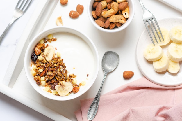 Yoghurt met muesli, banaan, noten en rozijnen en een roze servet op een wit dienblad