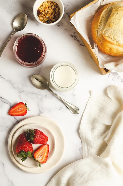 Yoghurt, vers fruit, aardbeienjam, muesli en brood met een wit servet op marmeren achtergrond.