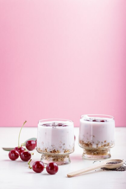 Yoghurt with cherries, chia seeds and granola in glass with wooden spoon on pink and white 