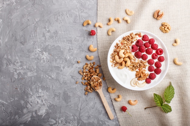 Yoghurt with raspberry, granola, cashew and walnut in white plate with wooden spoon on gray concrete background and linen textile. top view.