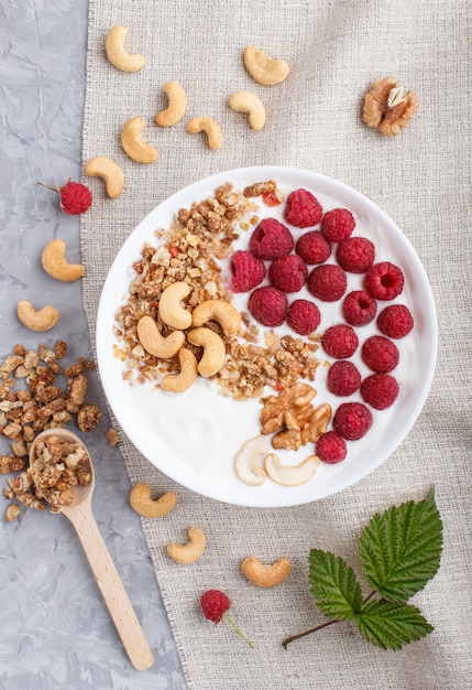Yoghurt with raspberry, granola, cashew and walnut in white plate with wooden spoon on gray concrete background