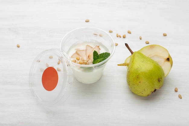 Ломтики йогурта, груши, мяты и кедровые орехи лежат в коробке для завтрака на белом столе рядом с разбросанными