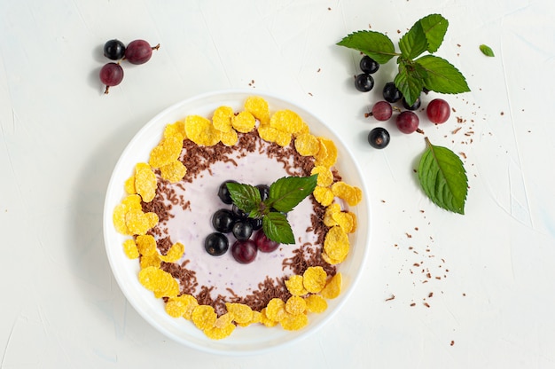 Yogurt with berry syrup decorated with chocolate currants, gooseberry and cornflakes and mint on a white concrete background. Copy spase. Top view.