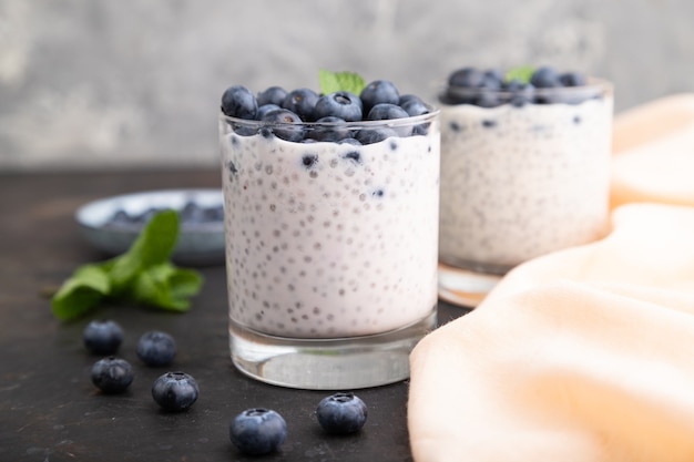 Yogurt with blueberry and chia in glass on black concrete surface and orange linen textile