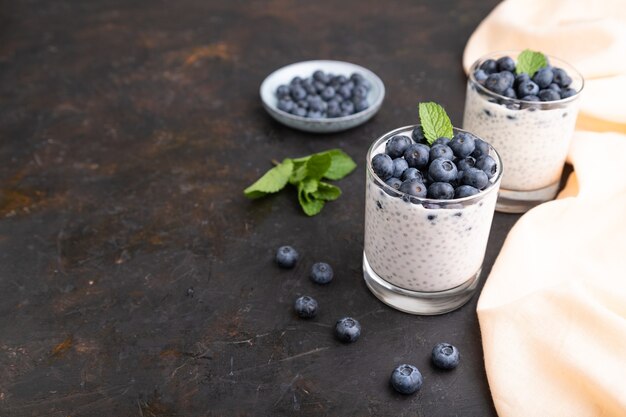 Yogurt with blueberry and chia in glass on black concrete surface and orange linen textile