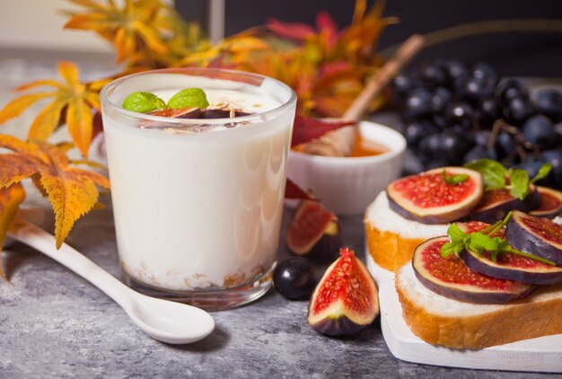 Yogurt with muesli and fig fruit in a glass, sandwich with cream cheese, figs and honey served on a small white cutting board. Healthy food . Autumn harvest.