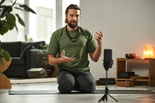 Yong man sitting on exercise mat in front of smartphone and communicating with his followers during