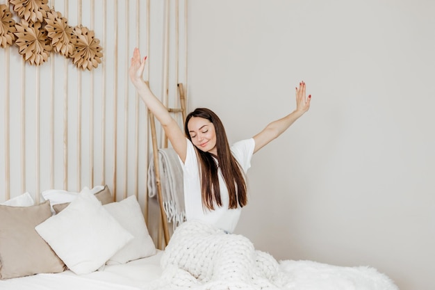 Yong woman sitting on the bed stretching after sleep and smiling in her room happy morning