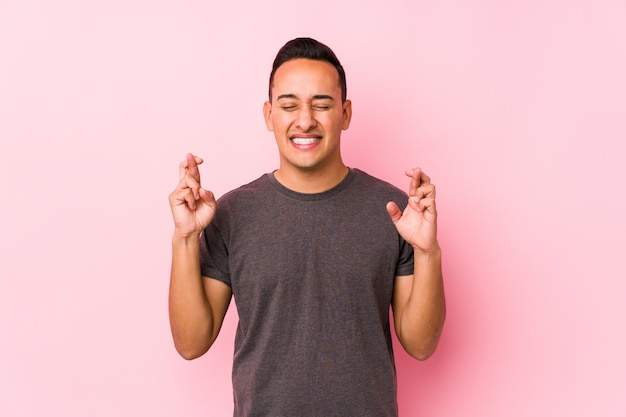 Молодой латинский мужчина позирует на розовом фоне скрещивая пальцы за удачу