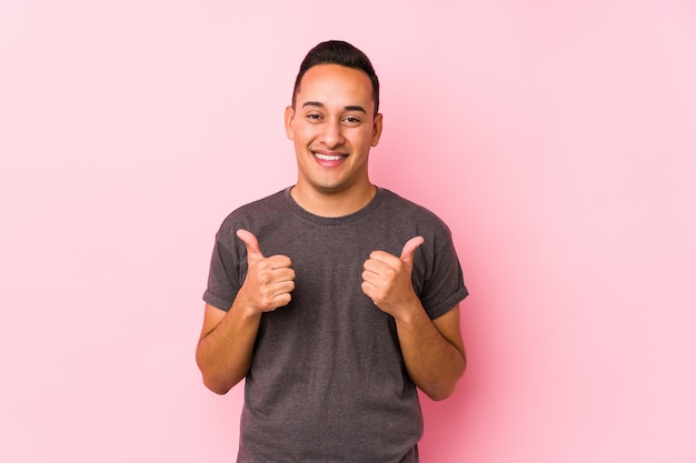 Yooung latin man posing in a pink wallraising both thumbs up, smiling and confident.