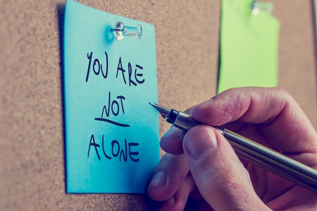 Photo you are not alone - man writing an inspirational message on a blue sticky note pinned to a cork board with a fountain pen.