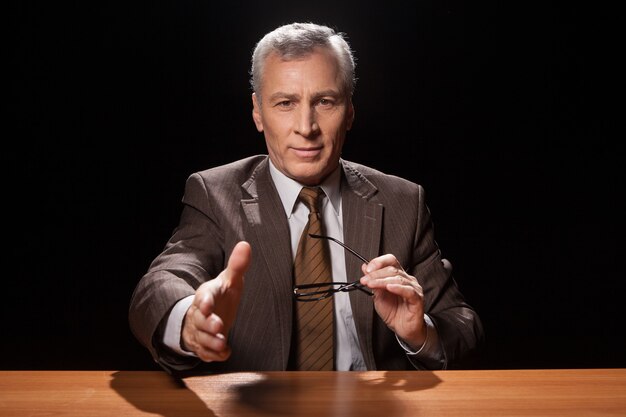 You are hired! Cheerful senior man in formalwear sitting at his working place and stretching out his hand for shaking while isolated on black background