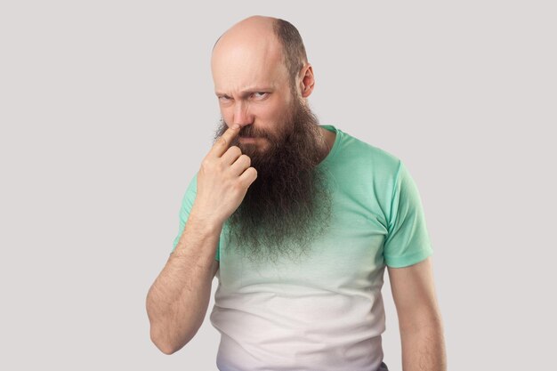 Photo you are liar portrait of angry middle aged bald man with long beard in green tshirt standing looking and pointing at his nose showing lie gesture indoor studio shot isolated on grey background