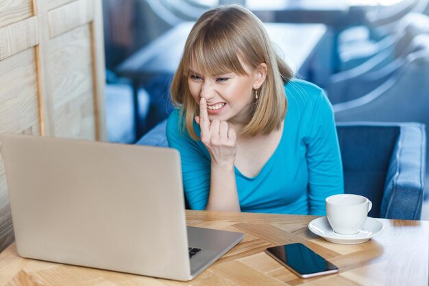 You are liar. Portrait of angry young girl freelancer with blonde hair in blue t-shirt are sitting in cafe and making video call on laptop, talking about dishonest and angry, pointing finger to nose.