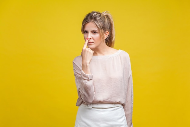 You are liar Portrait of dishonest young woman with fair hair in casual beige blouse standing touching her nose with finger showing lie gesture indoor studio shot isolated on yellow background