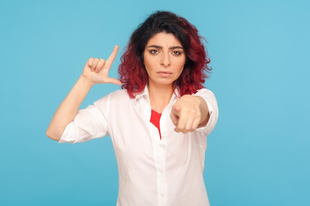 You are loser. Portrait of angry woman with fancy red hair showing L sign and pointing finger to camera, doing loser gesture to blame for failure. indoor studio shot isolated on blue background