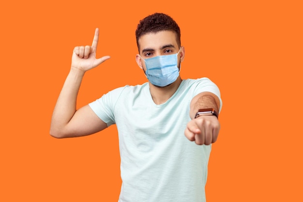 You are loser Portrait of bossy angry man with medical mask in white tshirt showing loser gesture and pointing at camera mocking your defeat indoor studio shot isolated on orange background