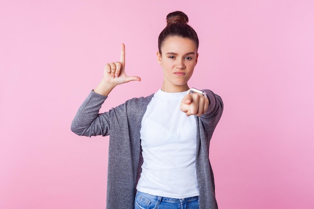 You are loser Portrait of brunette teenage girl with bun hairstyle in casual clothes showing loser gesture and pointing at camera blaming for failure indoor studio shot isolated on pink background