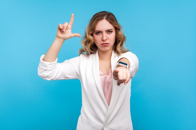 You are loser. Portrait of irritated woman with wavy hair in white jacket doing loser gesture, L finger sign and pointing at camera, blaming for failures. studio shot isolated on blue background