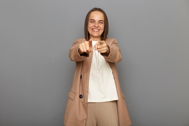 You are special Positive happy female wearing beige jacket standing pointing fingers at camera and looking with toothy smile believes in you motivation posing isolated over gray background