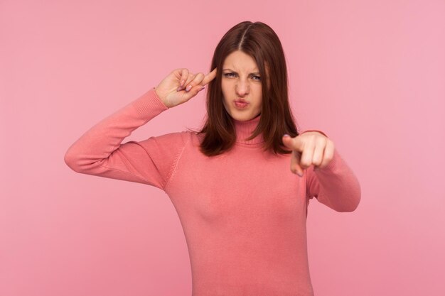 You are stupid Sad dissatisfied brunette woman showing stupid gesture pointing finger at you looking at camera blaming and mocking Indoor studio shot isolated on pink background