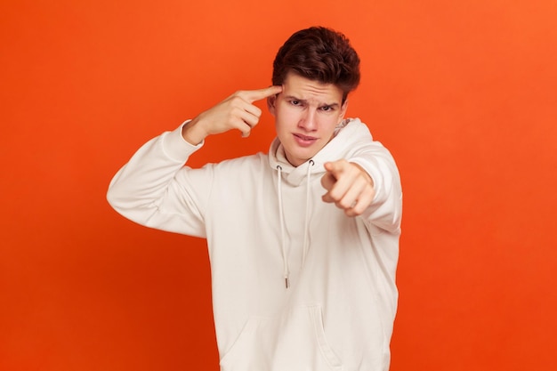 You are stupid. Self confident egoistic teenager holding finger near temple and pointing on you, showing stupid gesture, mocking. Indoor studio shot isolated on orange background