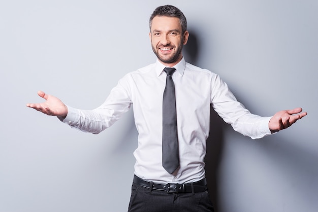 You are welcome! Cheerful mature man in shirt and tie gesturing welcome sign and smiling while standing against grey background