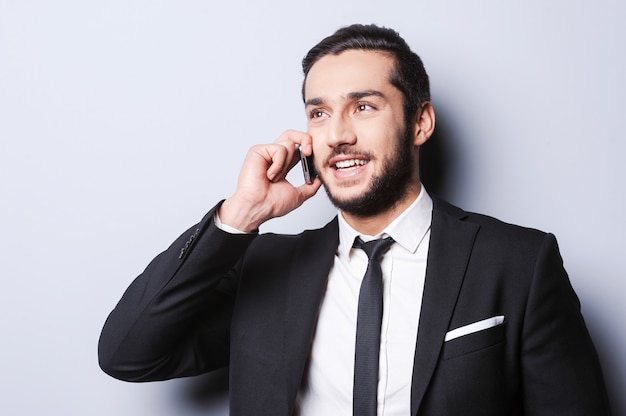 You can reach me anytime. Portrait of confident young man in formalwear talking on the mobile phone and smiling while standing against grey background