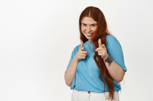 You got this, well done. Smiling redhead girl encourage you, boosting confidence, pointing fingers at camera to praise, compliment effort, standing against white background.