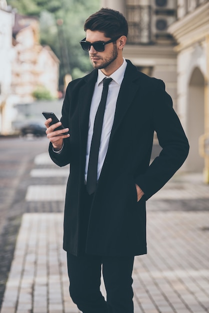 You have new message! Handsome young well-dressed man in sunglasses using his smart phone while walking outdoors