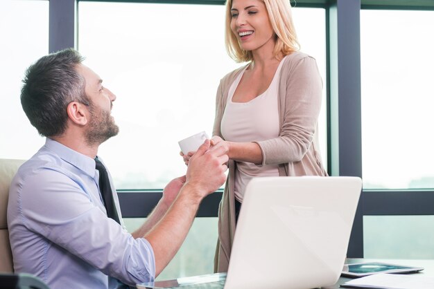 Foto hai bisogno di una pausa! donna allegra che dà una tazza di caffè a un uomo in abiti da cerimonia seduto al suo posto di lavoro