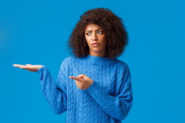 You see it awful. Unimpressed sad and displeased african-american woman having worst new year ever, pointing and presenting her presents with lack of interest, standing upset blue 