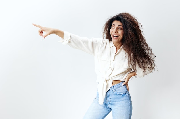 Do you see that portrait of amazed pretty curly latin woman\
staying near white studio wall background smiling look at the left\
and point finger at free place copy space for clothes sales