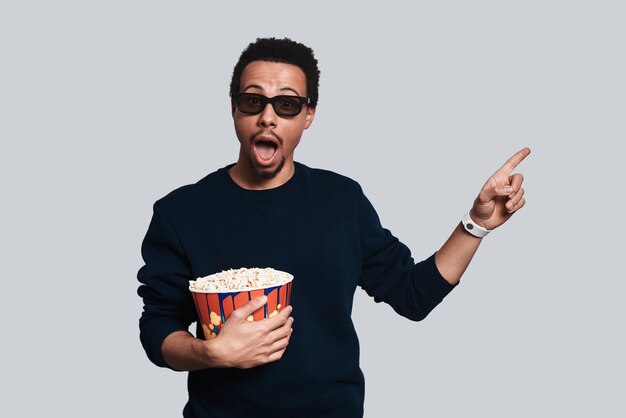 You should see it! Handsome young man in casual clothing looking at camera and pointing copy space while standing against grey background