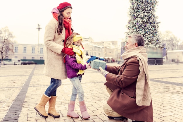 For you. Shy girl looking at box with present while standing close to her mother