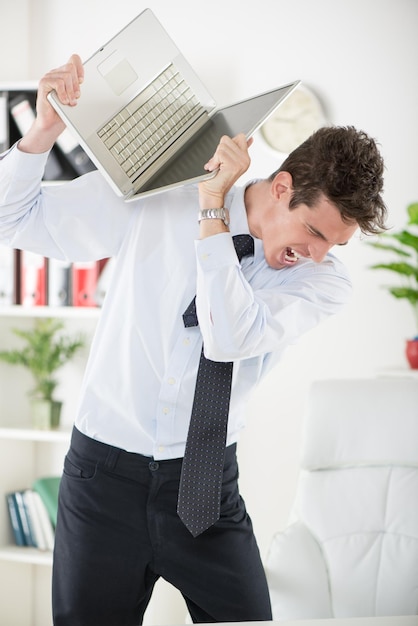 Youn uomo d'affari arrabbiato in piedi in ufficio e in possesso di un laptop come se volesse romperlo.