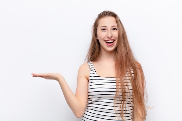 Yound blonde vrouw op zoek verrast en geschokt, met kaak gevallen met een object met een open hand aan de zijkant tegen de witte muur