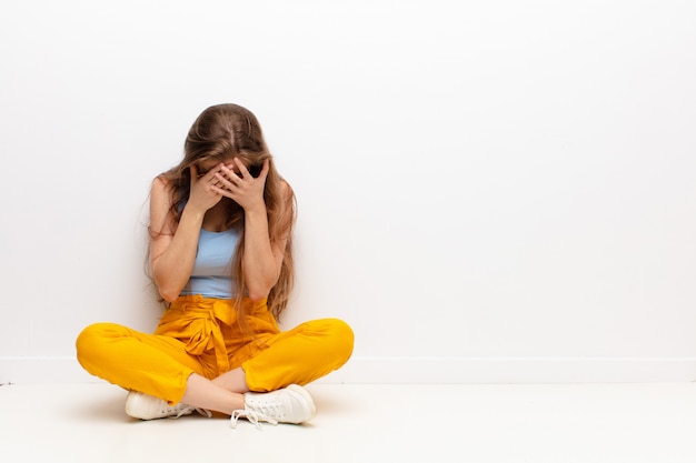 Yound blonde woman feeling sad, frustrated, nervous and depressed, covering face with both hands, crying sitting on the floor