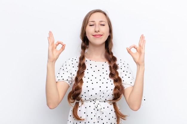 Yound blonde woman looking concentrated and meditating, feeling satisfied and relaxed, thinking or making a choice against white wall