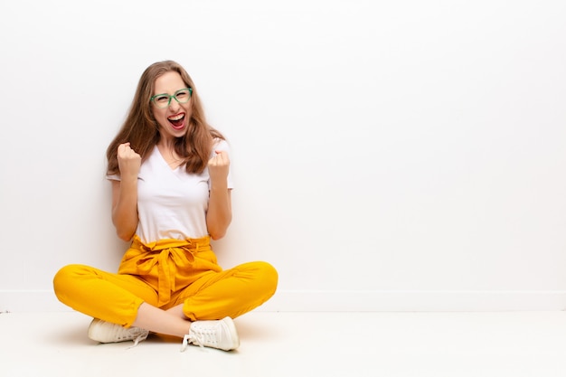 Yound blonde woman shouting triumphantly, laughing and feeling happy and excited while celebrating success sitting on the floor