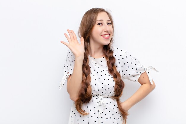 Yound blonde woman smiling happily and cheerfully, waving hand, welcoming and greeting you, or saying goodbye over white wall