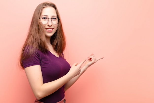 Yound blonde woman smiling happily and pointing to side and upwards with both hands showing object in copy space