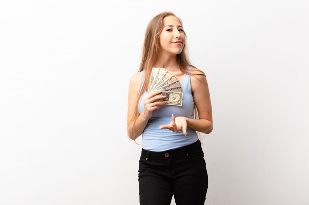 Photo yound blonde woman smiling happily with friendly, confident, positive look, offering and showing an object or concept holding dollar banknotes