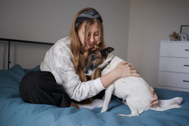 A young 20s girl sits on a bed in her living room and hugs a small white dog