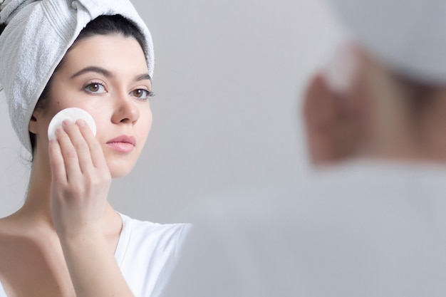 Young 25-year-old woman caring of her skin on the face, standing near mirror