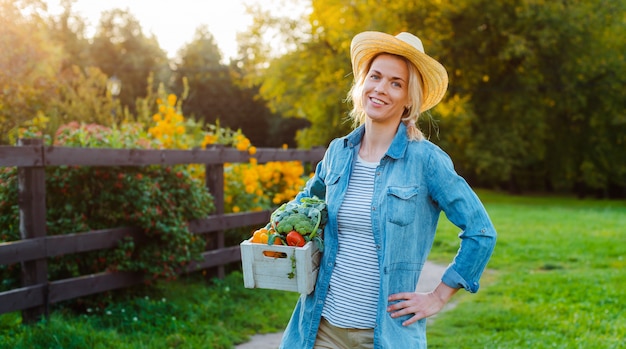 Молодая 30-35 лет красивая женщина-фермер в шляпе с коробкой свежих экологически чистых овощей