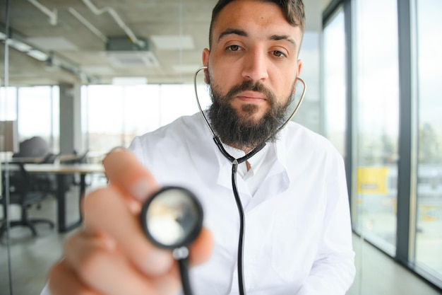 Giovane medico maschio arabo bello piacevole di 30 anni in camice bianco che posa alla macchina fotografica all'interno