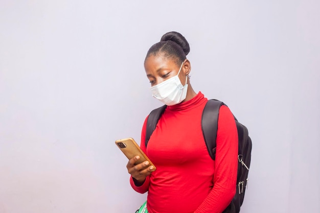 Молодая очаровательная чернокожая студентка в маске для носа с рюкзаком с помощью мобильного телефона
