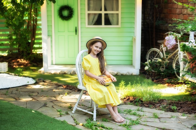 Young adorable girl sit on chair with apricots in colander at summer backyard at home